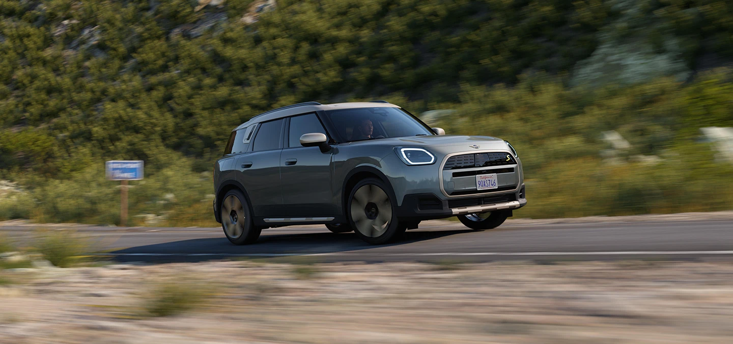 Side view of a driving all-electric MINI Countryman in Smokey Green with Vibrant Silver Roof offroad.